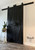Two tall, black sliding barn doors with black handles are mounted on a wall. To the left, a potted plant sits on a small stand above three white candles. The floor is wood, and the “Theresa Lee NTS Design Co.” watermark is on the wall.