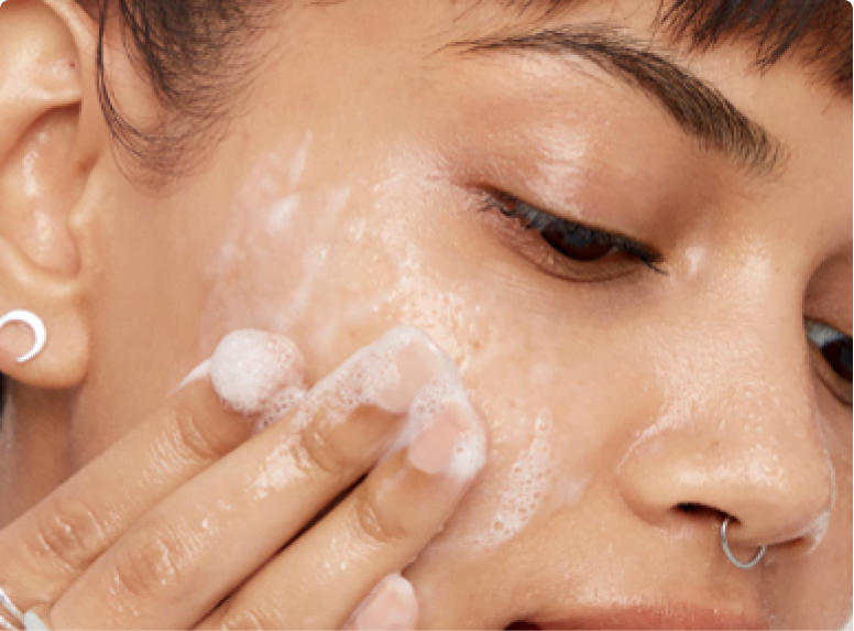 Female model with dark hair, nose ring, and crescent moon earring. She is cleaning her face with a foaming cleanser as step 1 in her skin care routine