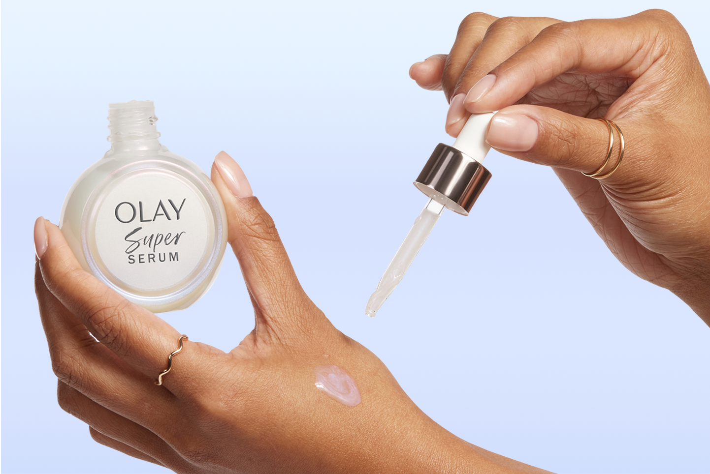  A close-up of a woman’s hands where her left hand is holding a bottle of the Olay Super Serum while her right hand holds its dropper to squeeze a bit of serum on the back of her left hand.