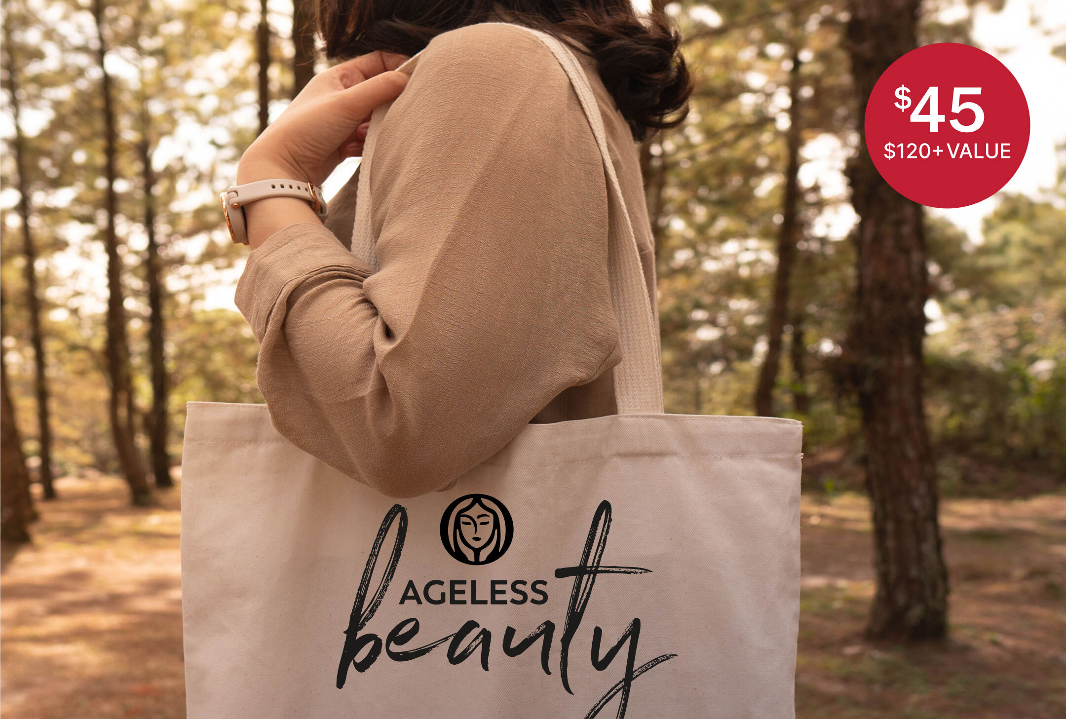 Person carrying a neutral canvas tote bag that has the Olay logo with a printed graphic that reads “Ageless Beauty” in a wooded outdoor setting. A red price badge reads $45 with $120+ value