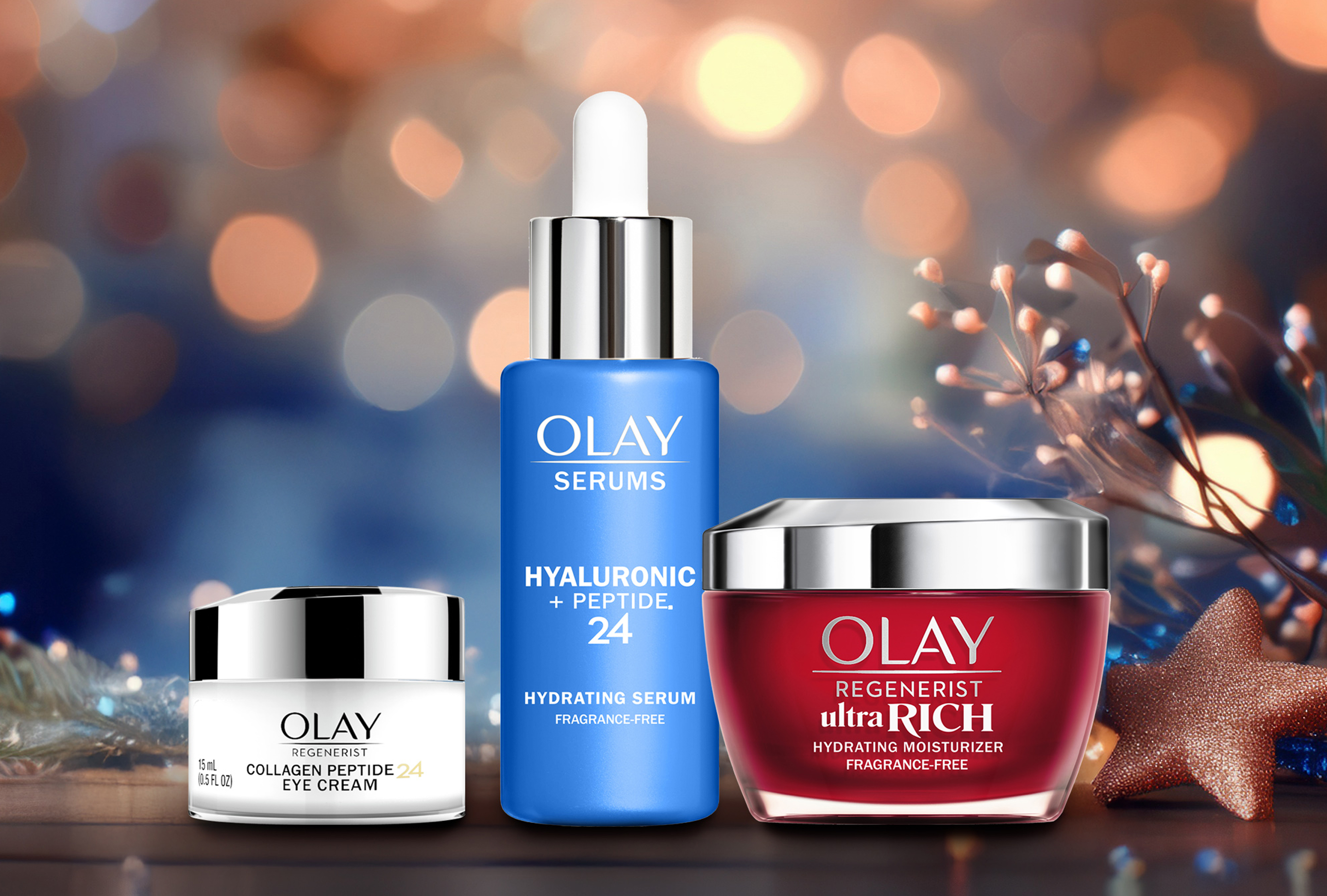 Olay Regenerist Ultra Rich Hydrating Moisturizer (red jar), Olay Serums Hyaluronic + Peptide 24 (blue pump), and Olay Regenerist Collagen Peptide 24 Eye Cream (white jar) arranged on a brown table with festive decorations and soft light in the background.