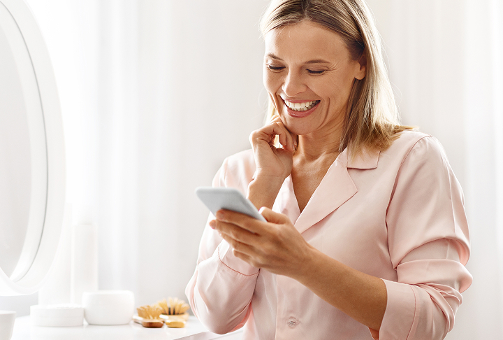 Woman wearing light pink pajamas sitting beside her bedroom vanity mirror looking at her cellphone and smiling.