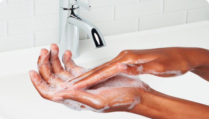 Une personne se frottant les mains avec un nettoyant mousseux sous un robinet qui coule dans une salle de bains carrelée blanche.