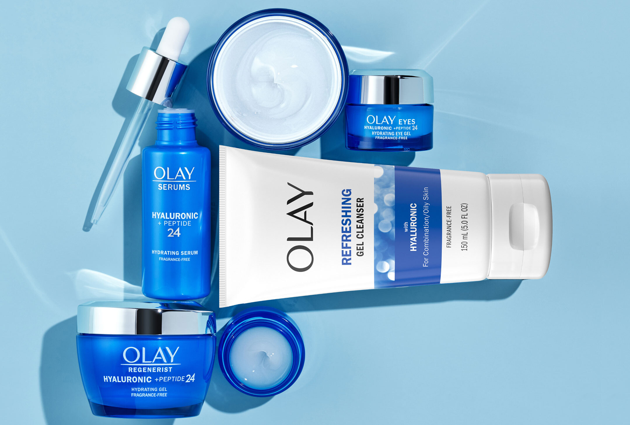 Olay Hyaluronic + Peptide 24 skincare line including cleanser, serum, gel cream, and eye gel arranged flat on a blue background.