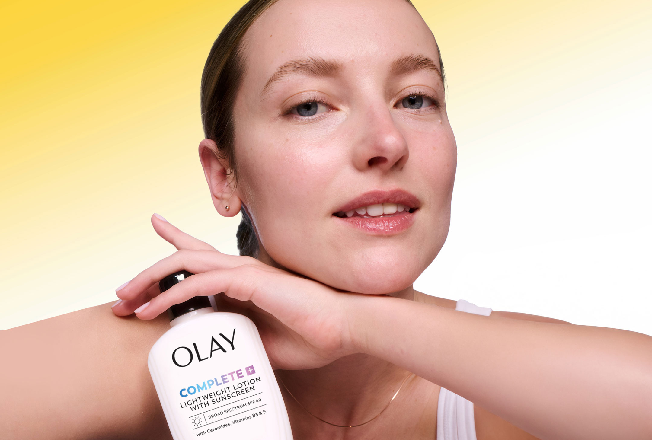 Smiling woman with dewy, natural skin holding a bottle of Olay Complete Lightweight Lotion with SPF 30 against her cheek, set against a yellow-to-white gradient background resembling a sunny day.