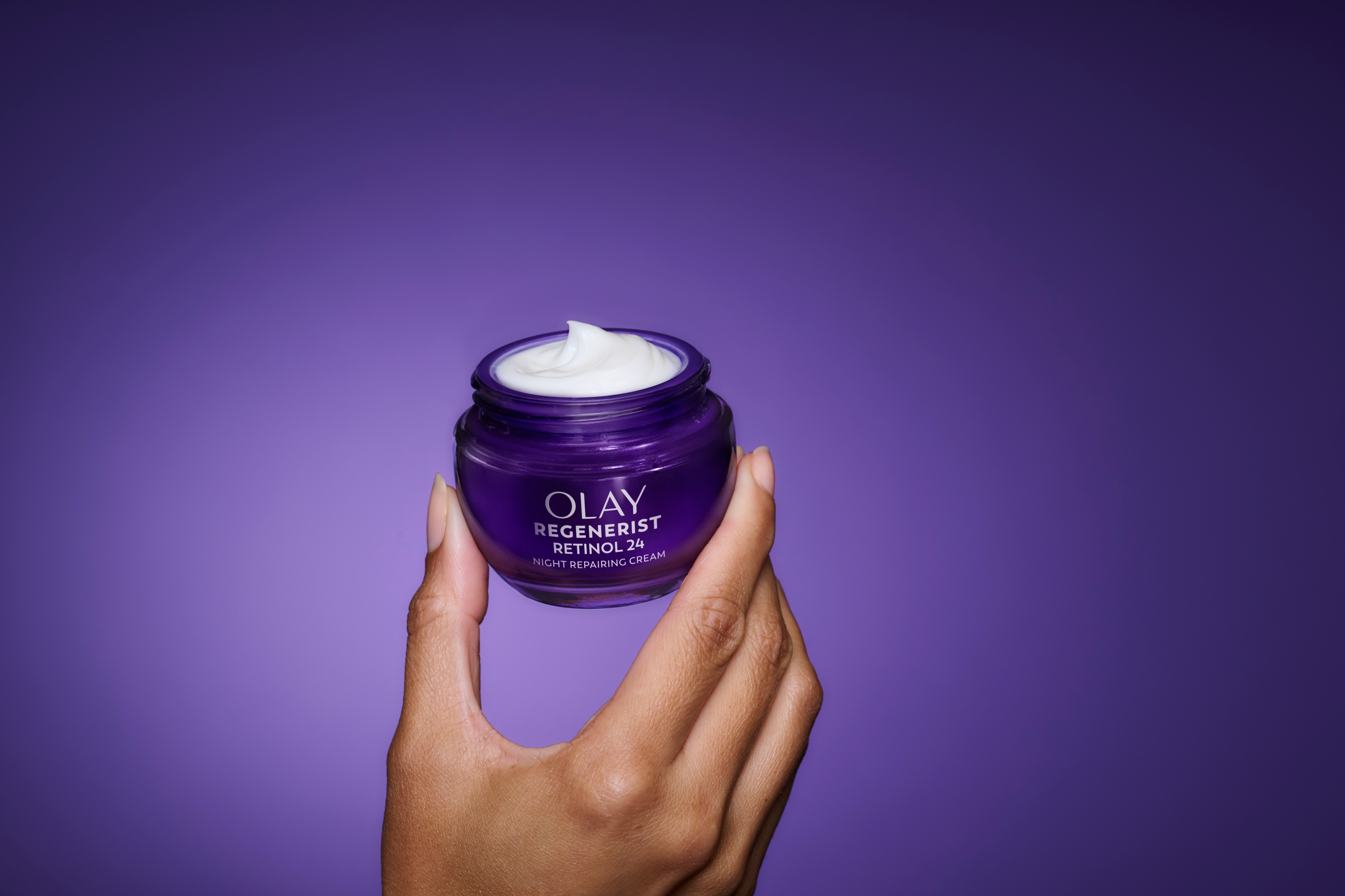 Deep purple glass jar of Olay Retinol24 Night Face Moisturizer with its lid removed and held up by a woman’s hand to lift it into a spotlight