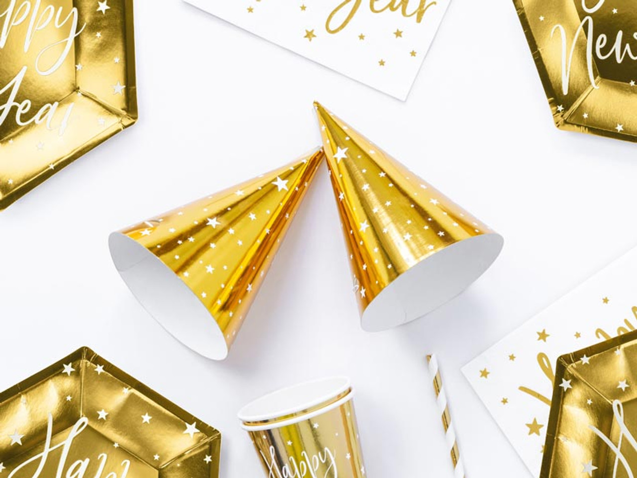 Two gold party hats, a cup, a straw, and festive plates are arranged on a white background.