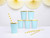 A stack of light blue paper cups with gold scalloped edges, accompanied by striped yellow and white straws, set against a pastel backdrop.