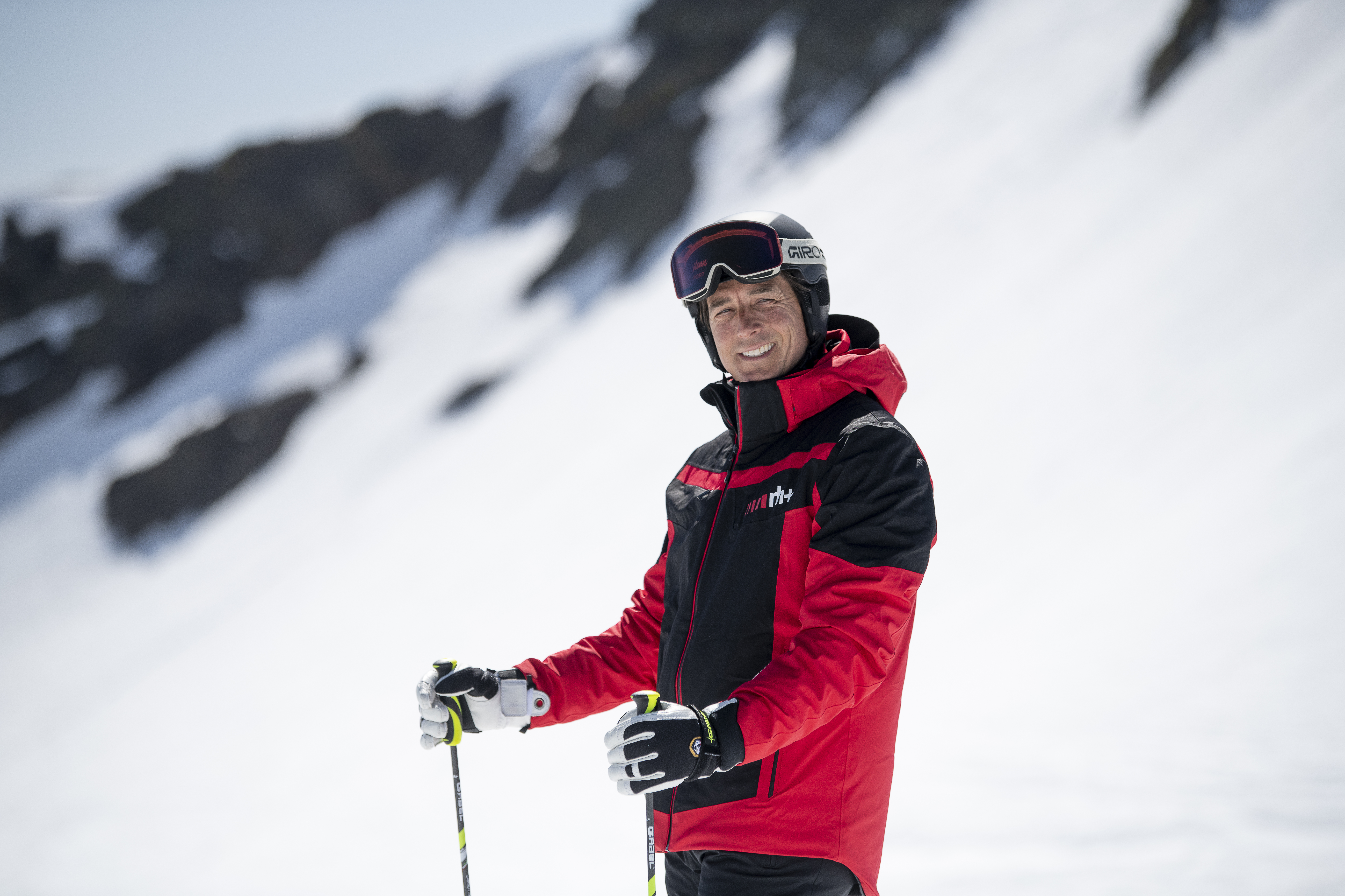 man smiling wearing ski jacket, goggles, and helmet, holding ski poles