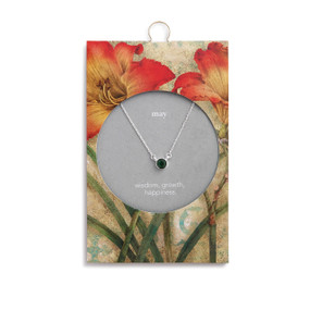 Silver chain necklace holds an emerald pendant. Packaging has yellow and red lilies