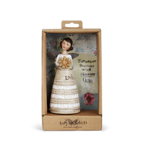 Angel figurine with brown hair and wings wears a dress with white top and sand&white striped skirt. Brown engraved lettering on skirt. Angel holds a yellow flower. Angel is in brown gift packaging