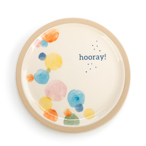 An ivory circular ceramic plate with a gold painted rim, multicolored watercolor painted dots, and hooray!" surrounded by small black dots."