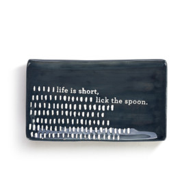 A ceramic, black, rectangular spoon rest with black rectangular dots, and reads life is short, lick the spoon"."