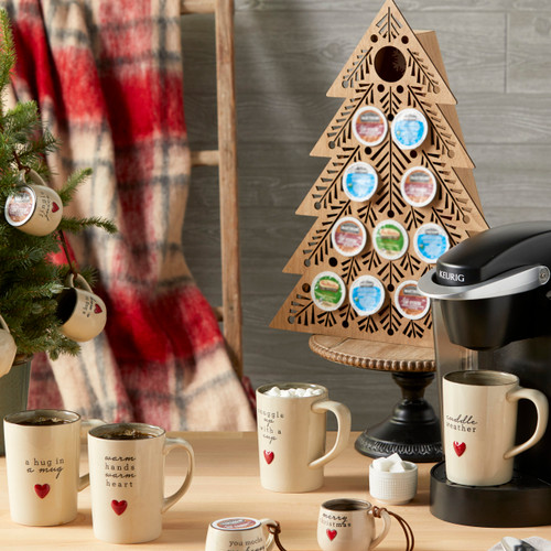 cream colored mugs and coffee pod holders with Christmas sentiments and red hearts on them sitting on a table and being used with a coffee with wooden tree shaped stand holding coffee pods