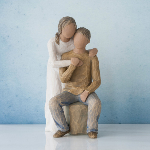 Man figurine sitting on rock wearing brown shirt and blue jeans with woman figurine in white dress with arms wrapped around him - blue background