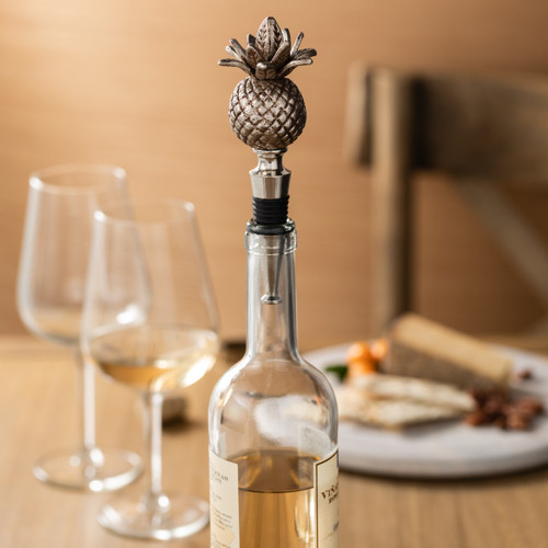 bottle stopper topped silver carving of pineapple in wine bottle sitting on table in front of wine glasses and cheese plate