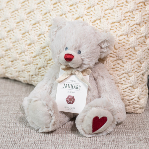 A cream plush birthstone bear with a dark red nose and embroidered heart on its foot for the January birthstone of Garnet, displayed sitting on a couch.