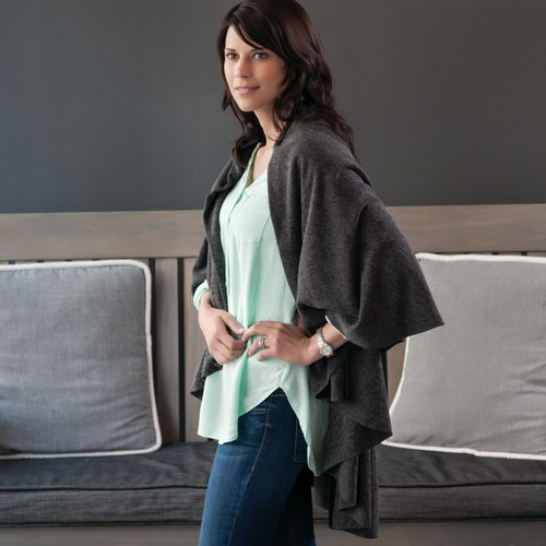 Side profile of dark haired model wearing dark grey cardigan draped over light blue shirt and blue jeans - grey bench in the background