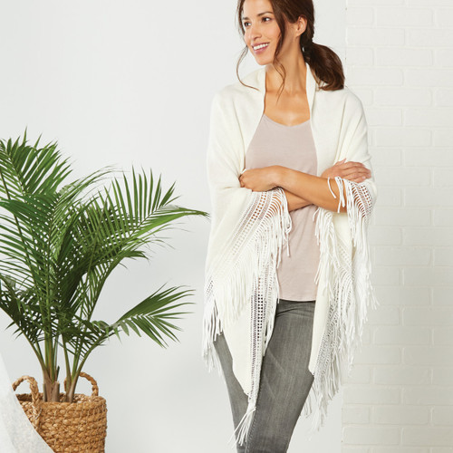 Far front view of brunette woman standing in white shawl with light pink shirt and grey jeans, plant visible in the background