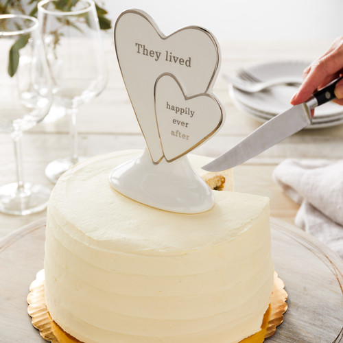 ceramic cake topper with two hearts with gold outline reading They lived Happily ever after on top of wedding cake while someone's cutting it