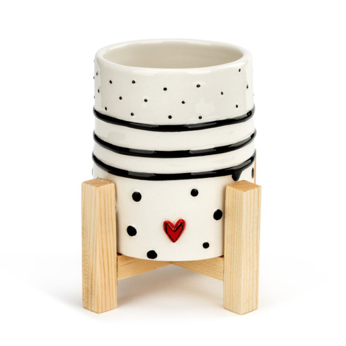 A small white planter with black polka dots, black stripes, and a small red heart. Placed in a light wooden stand.