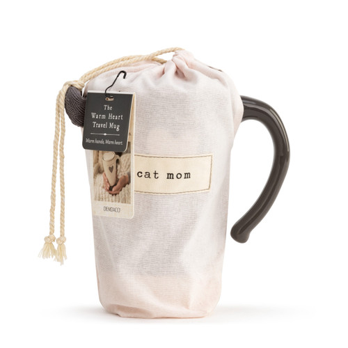 A tapered, charcoal, ceramic travel coffee mug, with a thick cream stripe base, a single cream heart in the center, and reads cat mom" in a cream typewriter font. With a handle, and lid placed on top. Placed inside a light pink drawstring bag that reads "cat mom" in an ivory patch, and has a product tag."