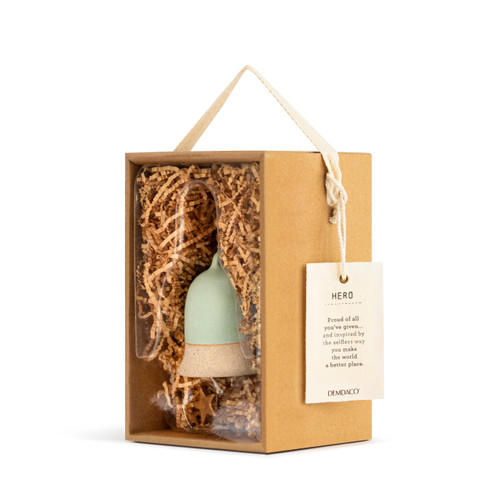 A left facing view of a mint green Inspired Bell" with a tan base, a twine rope, and gold and wooden beads. Placed in a brown cardboard box with brown crinkle paper, and an ivory tag that reads "Hero"."