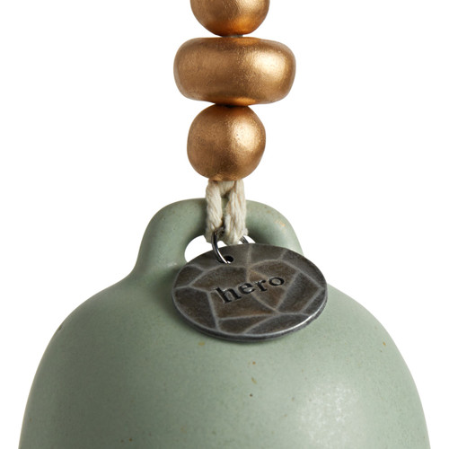 A close up image of the silver circular pendant that reads "hero" on a light green Inspired Bell.