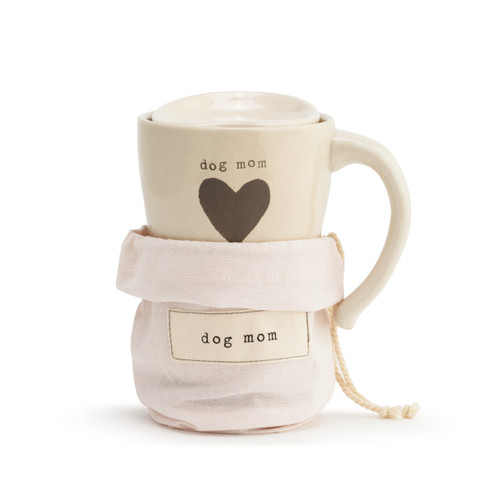 A tapered, cream, ceramic travel coffee mug, with a thick gray stripe base, a single gray heart in the center, and reads dog mom" in a gray typewriter font. With a handle, and lid placed on top. Halfway placed inside a light pink drawstring bag that reads "dog mom" in an ivory patch, and has a product tag."