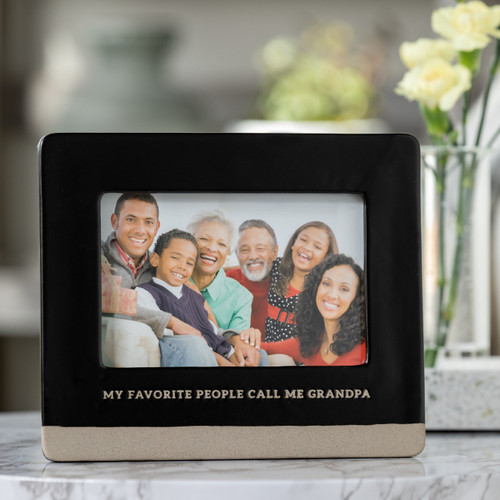 A close up image of a black and tan rectangular picture frame, engraved with my favorite people call me grandpa" and filled with a family photograph. Placed on a marble and wooden side table with a vase of flowers."