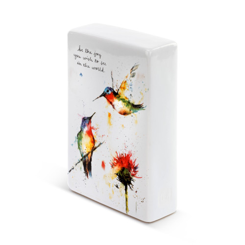 A side view of a white ceramic plaque with two painted hummingbirds flying above a red flower, and be the joy you wish to see in the world." in black cursive font."