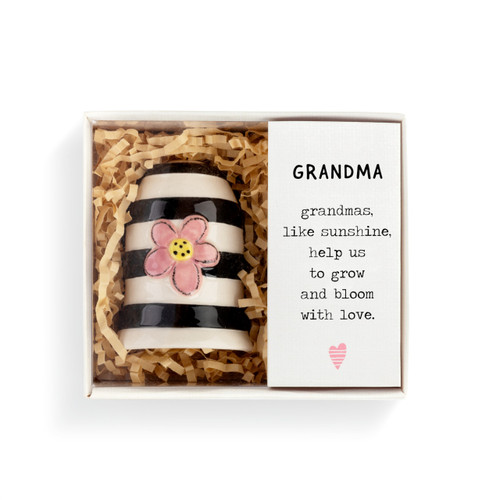 A black and white striped Heartful Home Bell" with a pink and yellow flower in the center, a chime that reads "Grandma", and a black ribbon string. Placed inside a white packaging box that reads "Grandma" and has brown crinkle paper."