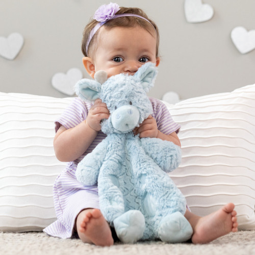 A close up image of a toddler girl sitting on a rug in front of large pillows holding a soft plush blue bear blankie.