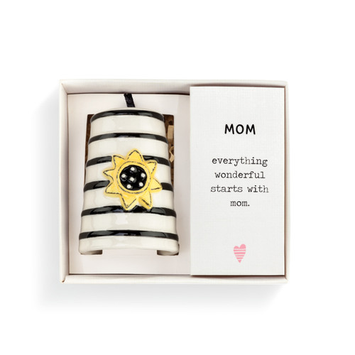 A white Heartful Home Bell" with black stripes, a yellow sunflower in the center, a chime that reads "friend", and a black ribbon string. Placed inside a white packaging box that reads "Friend" and has brown crinkle paper."