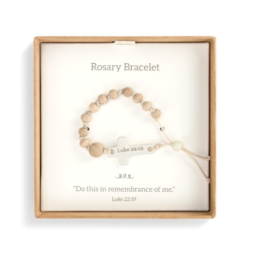 An ivory corded girl's rosary bracelet with several wooden beads, and a cream cross with Luke 22:19" engraved. Placed in a brown and white cardboard packaging that reads "Rosary Bracelet" and "Do this in remembrance of me" in beige font."