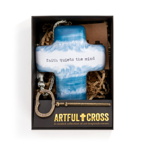 A blue and white art heart that reads faith quiets the mind" placed inside a black packaging box, with a bronze key, a silver tassel, and brown crinkle paper."