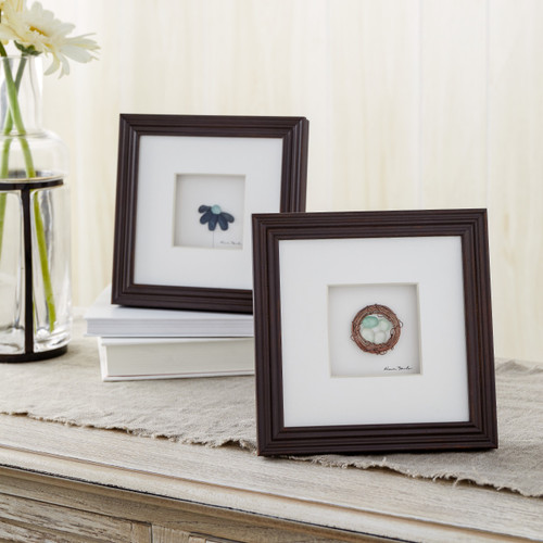 two dark wooden frames depicting a bird's nest and blue flower sitting on a dresser