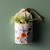 A cream ceramic magnet crock with an image of wildflowers and filled with greenery, attached to an olive green wall.