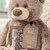A close up image of a plush brown plaid bear with a photo enclosed and a heart that reads "here to hug". Placed on a cream couch with a throw blanket.