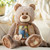 A close up image of a plush brown plaid bear with a father daughter photo enclosed and a heart that reads "here to hug". Placed on a cream couch with a throw blanket and pillow in a living room scene.