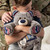 A close up image of a young boy hugging a gray and blue plush bear with a photograph in the center, and a heart that reads here to hug". In a living room setting."