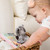 A close up image of a baby girl, reaching into a wicker basket beside an ivory couch in a living room scene. Looking at a small, plush, koala and a soft activity book called Hello! Around the world"."