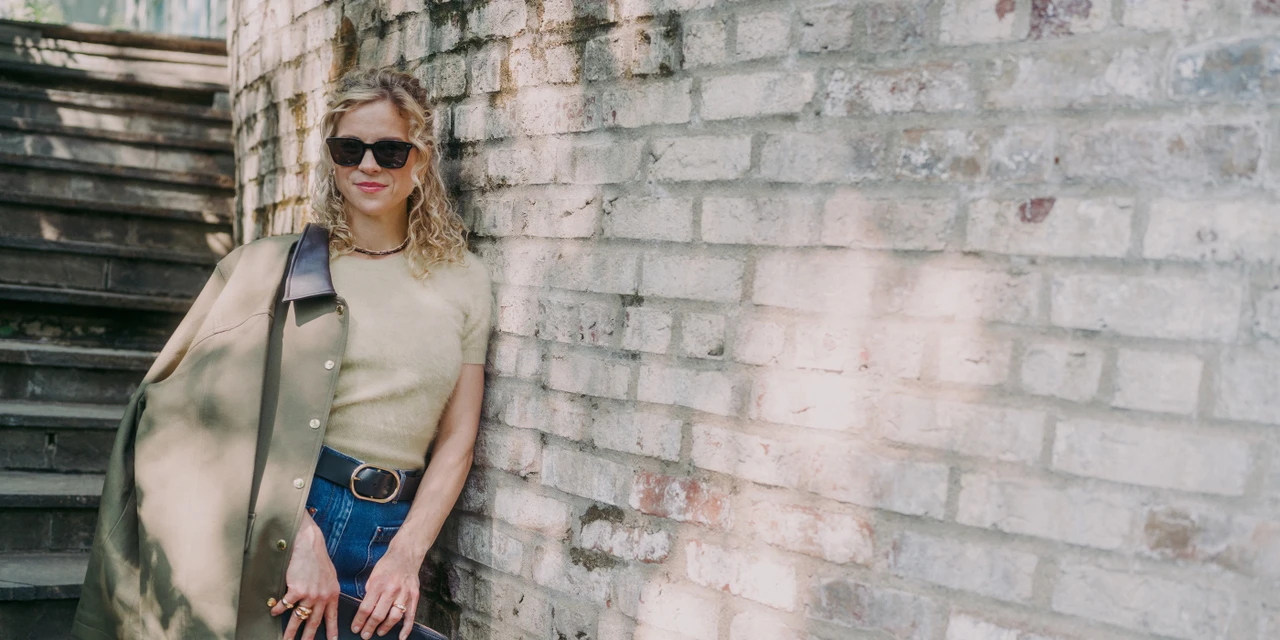 A woman outdoor in front of a brick wall with ivy, wearing a gray T-shirt, black hoodie, cross body bag and sunglasses talking on phone.