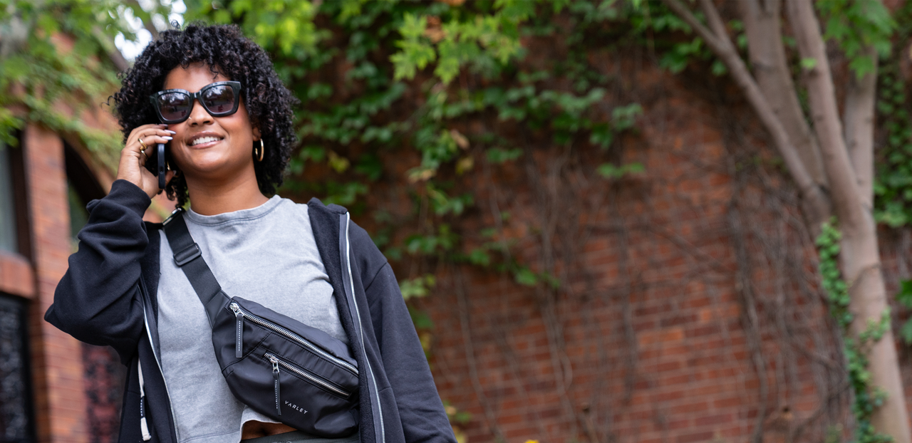 A woman outdoor in front of a brick wall with ivy, wearing a gray T-shirt, black hoodie, cross body bag and sunglasses talking on phone.