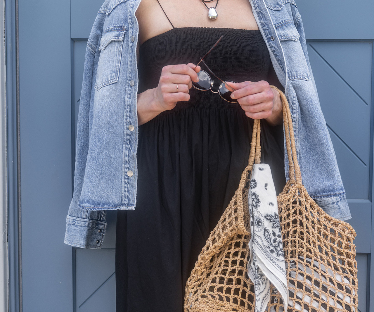 Person wearing an olive quilted jacket and black leggings, holding a large black leather tote bag with round sunglasses hooked on the handle.