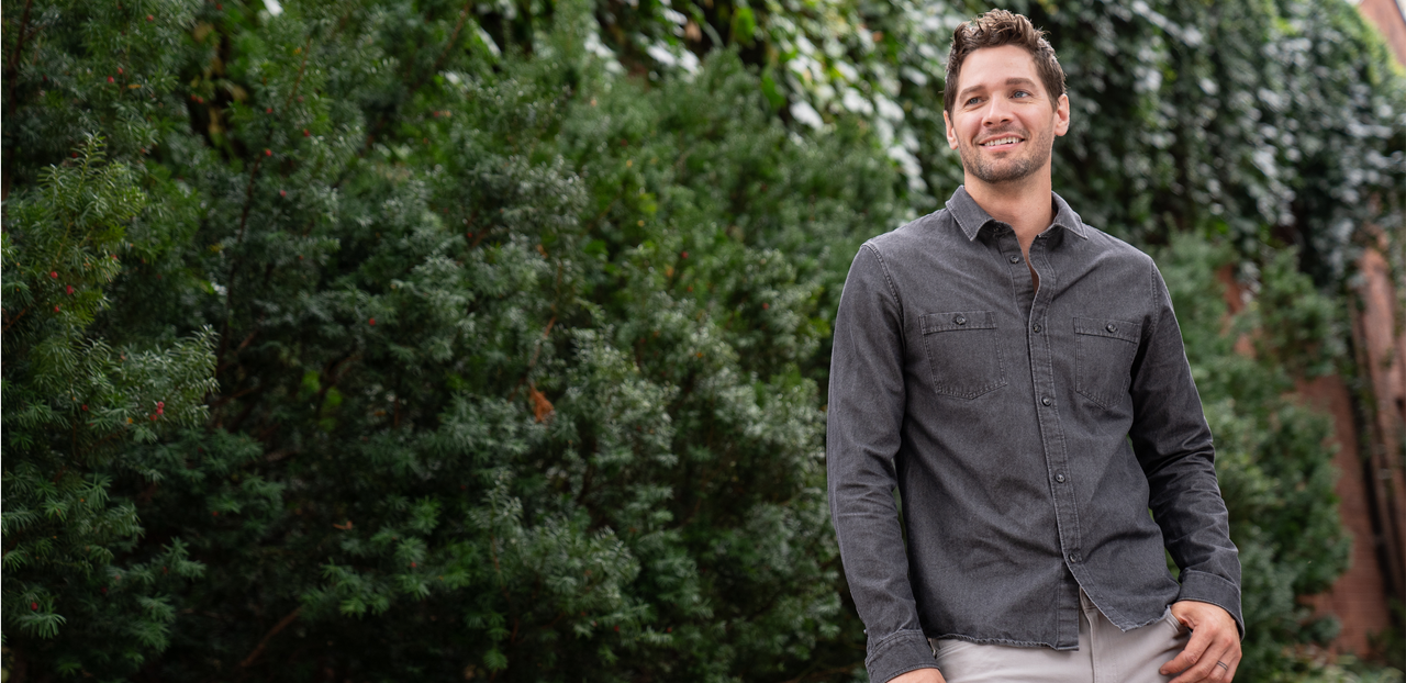 A man wearing a dark button-up shirt and light pants, standing outdoors in front of dense green foliage and ivy