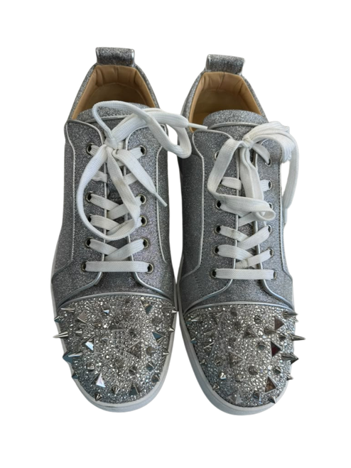 Vist Large View of Product Image Which is Christian Louboutin men’s silver glitter low-top sneakers with white laces, rhinestone and metallic spike embellished toe caps, and red soles.