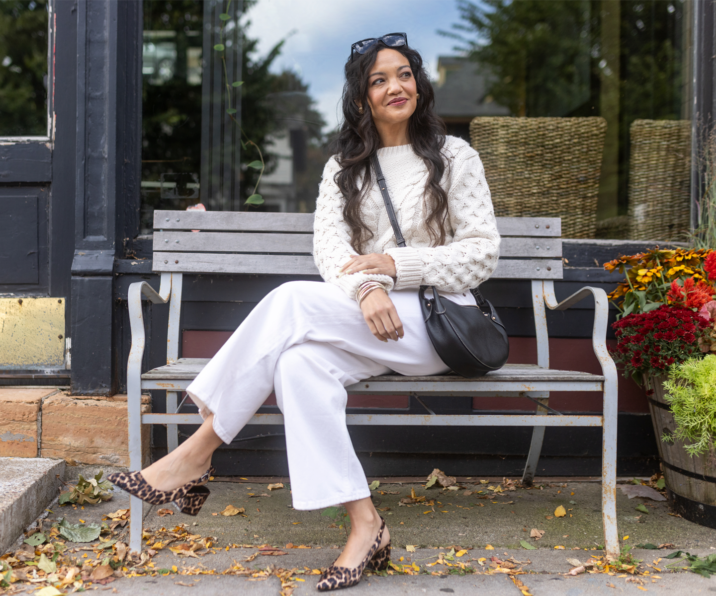 A woman sitting on a bench wearing a white knit sweater, white pants, leopard-print shoes, and a black crossbody bag.