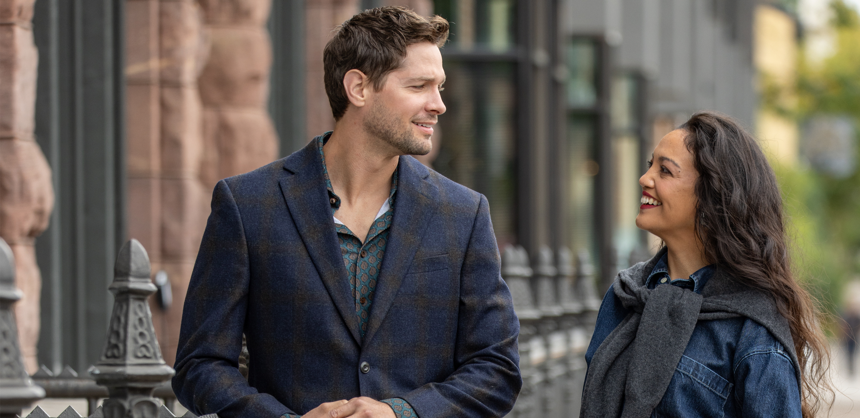A man and a woman standing outdoors near a metal fence, wearing dark jackets and talking on a city sidewalk.