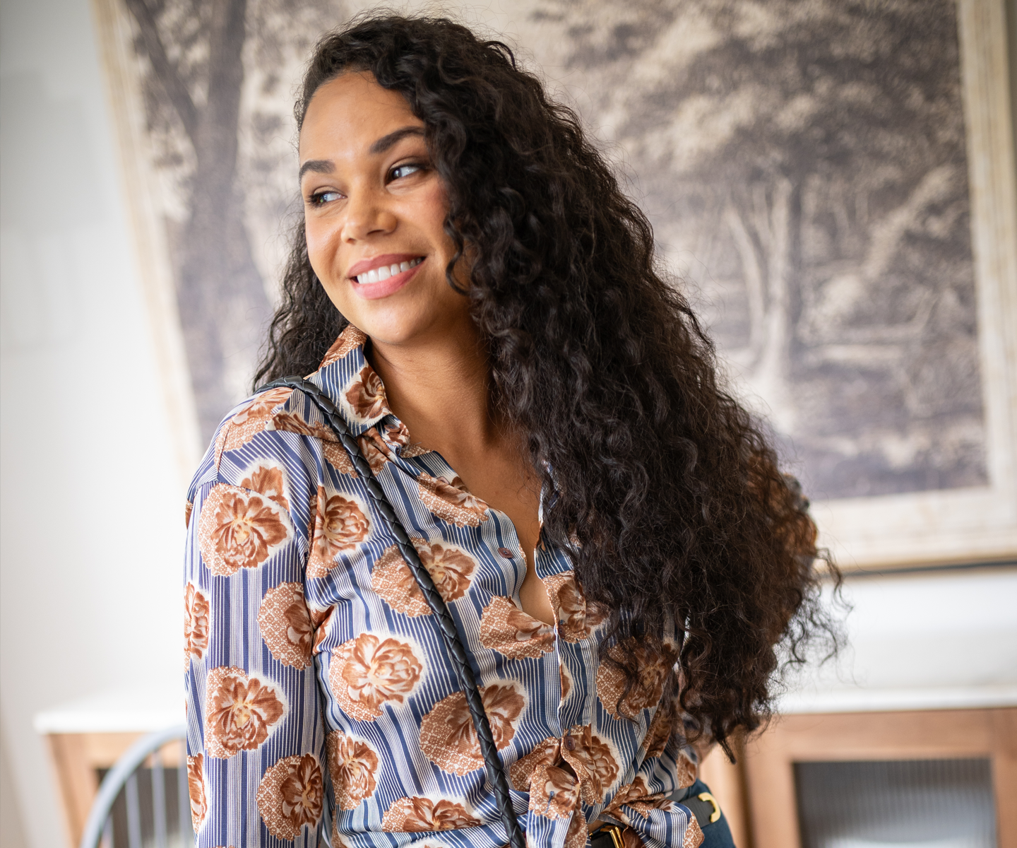 A woman wearing a patterned button-up shirt with large floral designs and a black crossbody bag indoors.